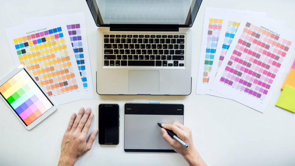 Eine Person verwendet ein Grafiktablett und einen Stift auf einem Schreibtisch mit einem Laptop, Farbmustern, einem Smartphone und einem Tablet – alles wichtige Tools zum Entwerfen einer nahtlosen Website-Navigation.